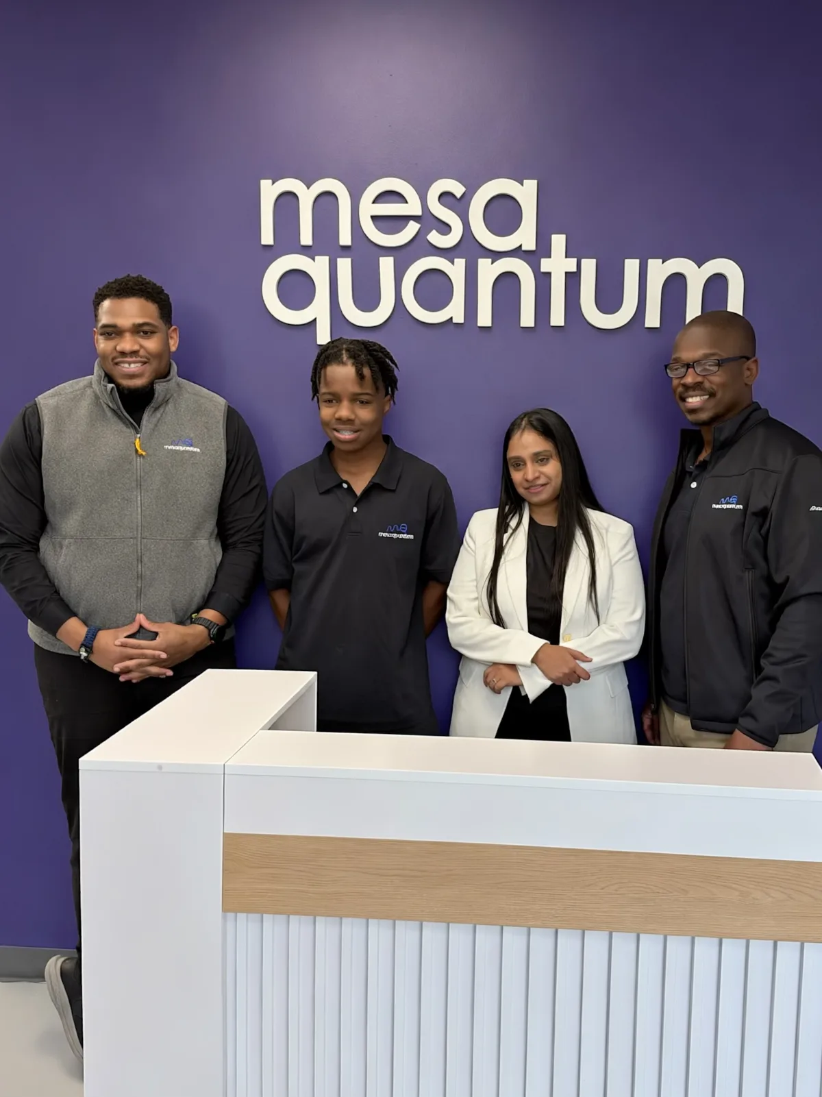 Titus, in a black Mesa Quantum polo, stands with TQ and two Mesa Quantum colleagues in front of the company's white wall logo at the reception desk.